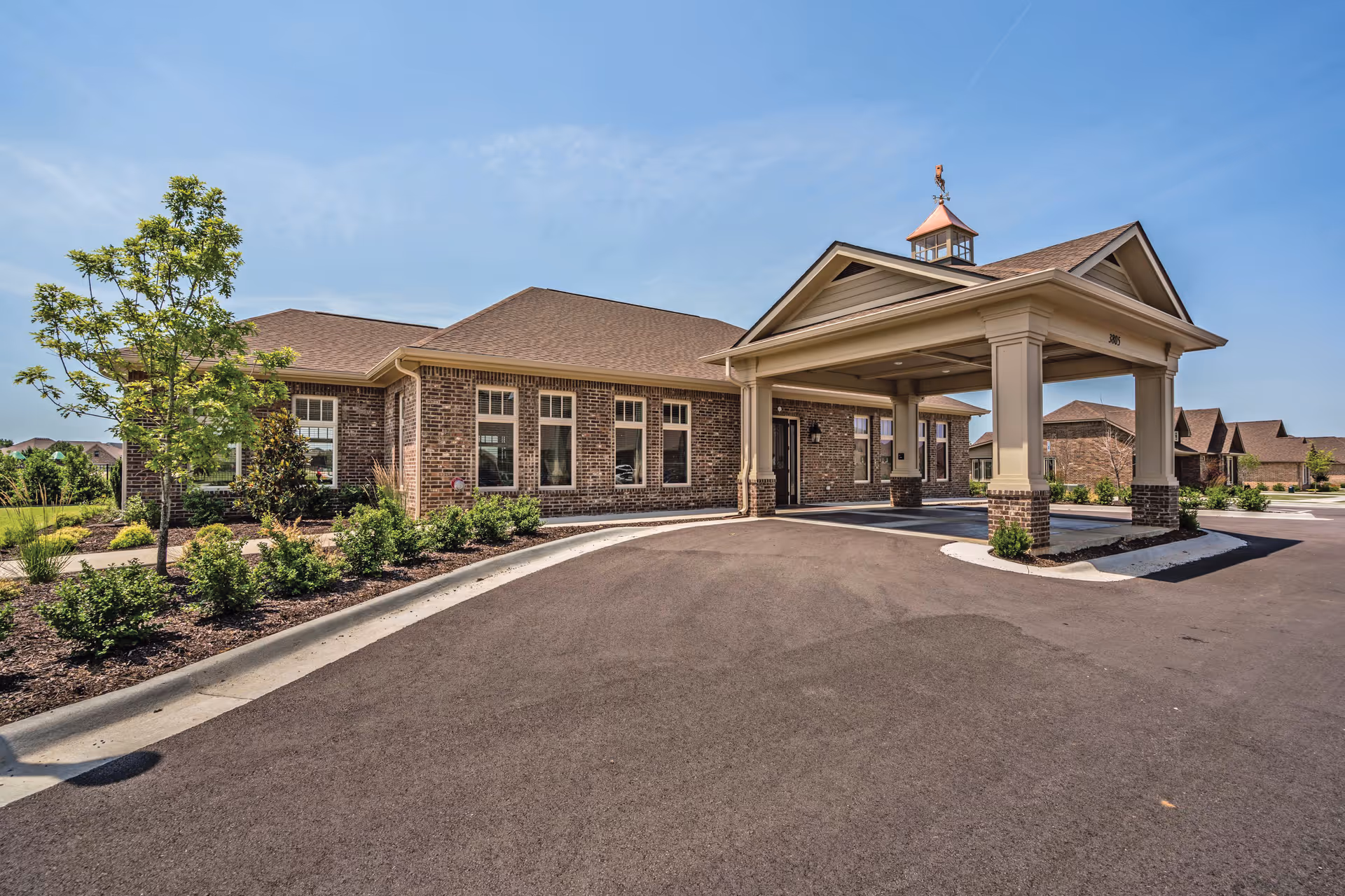 Exterior view of a single-story brick building with a covered entrance supported by columns. The building is surrounded by a paved driveway and landscaped with small bushes and a tree. The sky is clear and blue.
