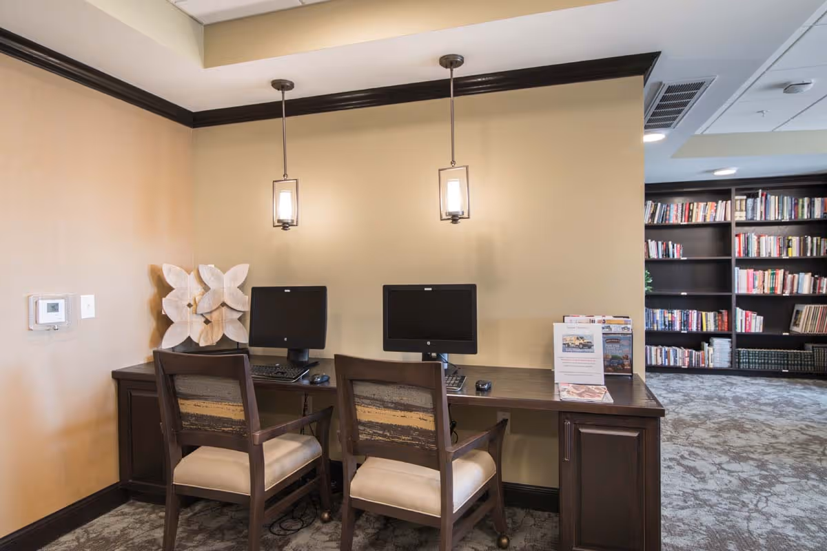 Two desktop computers and chairs on a wooden desk in a shared interior workspace next to bookshelves.