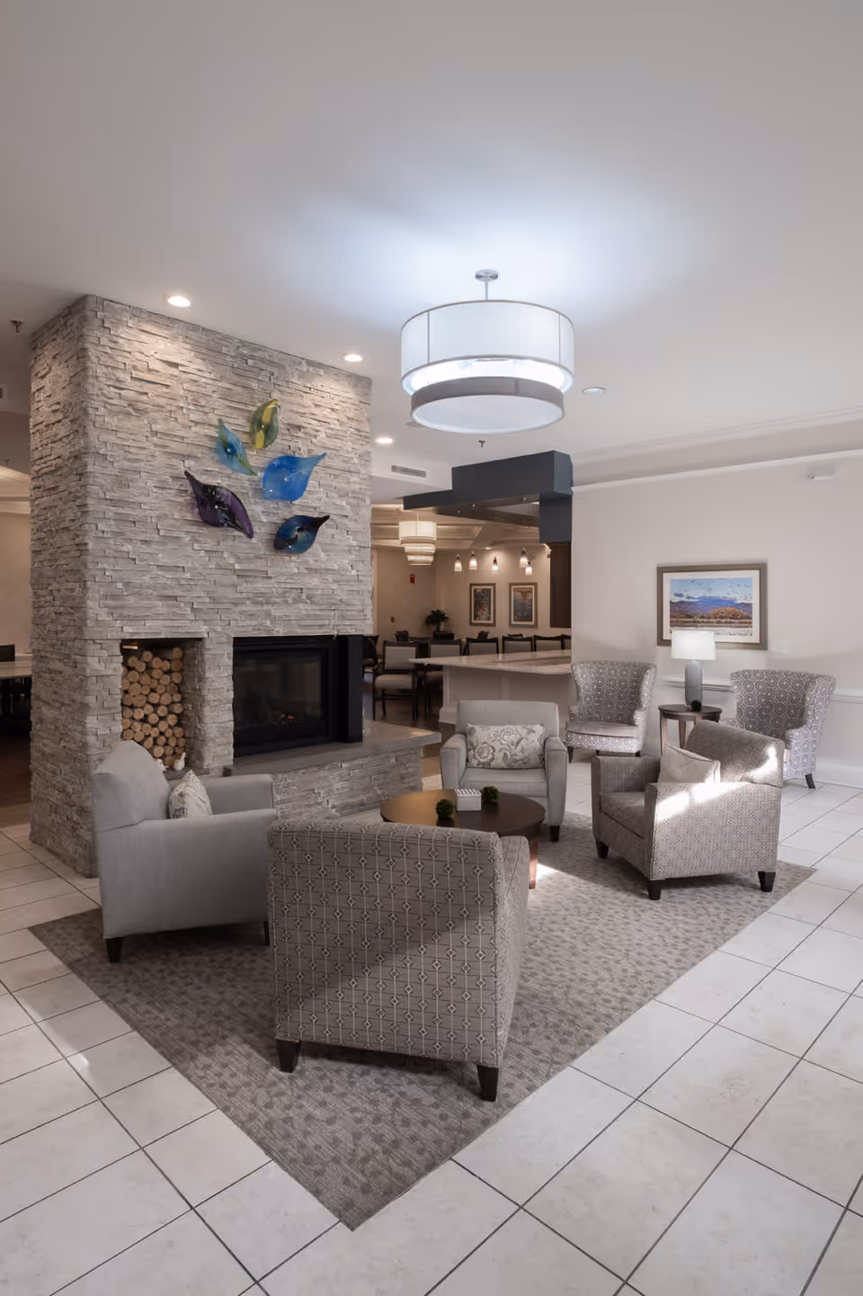 A cozy seating area in a senior living facility with four upholstered armchairs arranged around a round wooden coffee table on a patterned rug. The space features a stone fireplace with stacked firewood and colorful glass leaf decorations mounted above it. In the background, there is a dining area with tables and chairs, pendant lighting, and framed artwork on the walls.