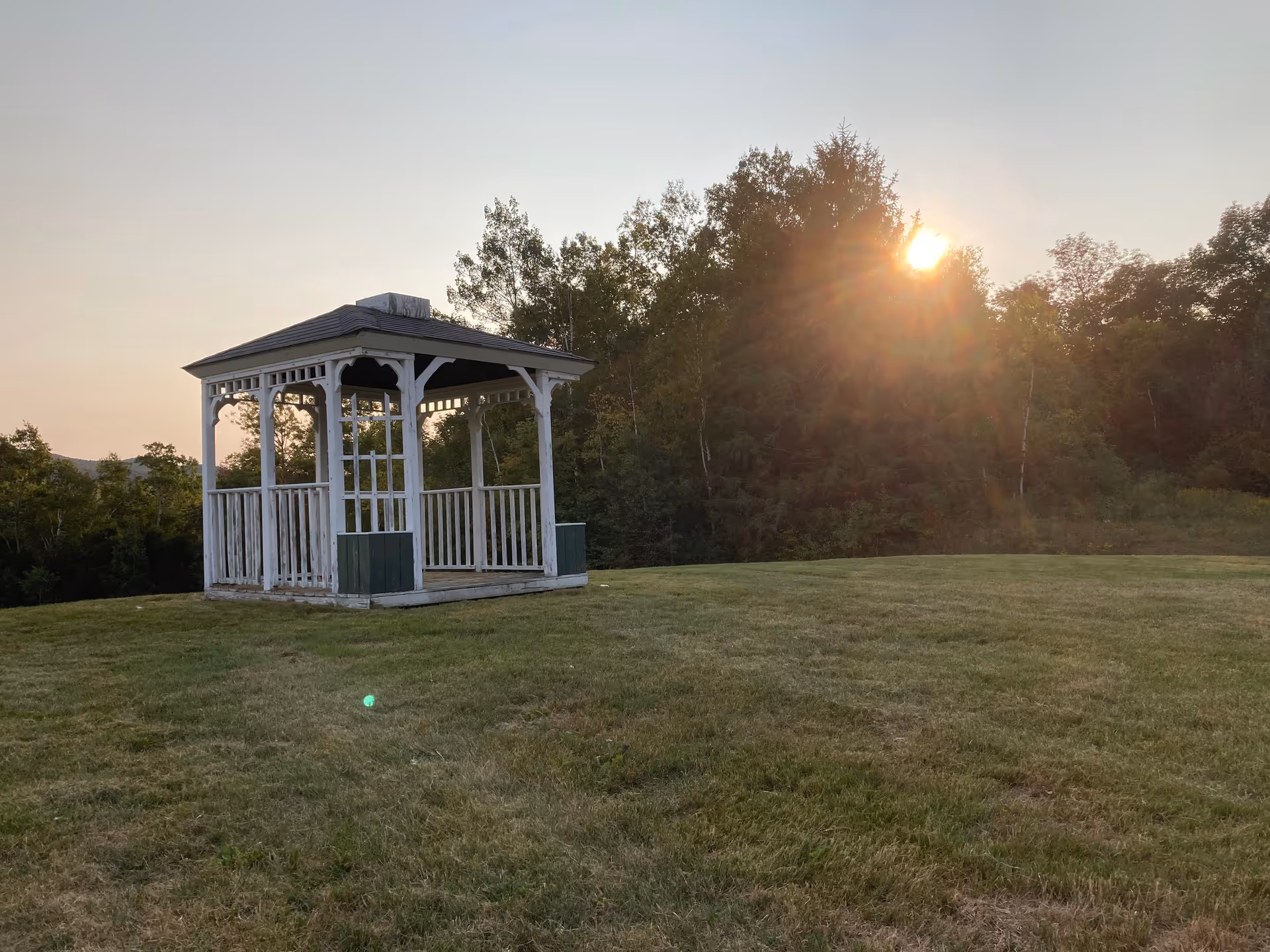 A white wooden gazebo standing on a grassy lawn with trees in the background and the sun shining through the trees during sunset or sunrise.