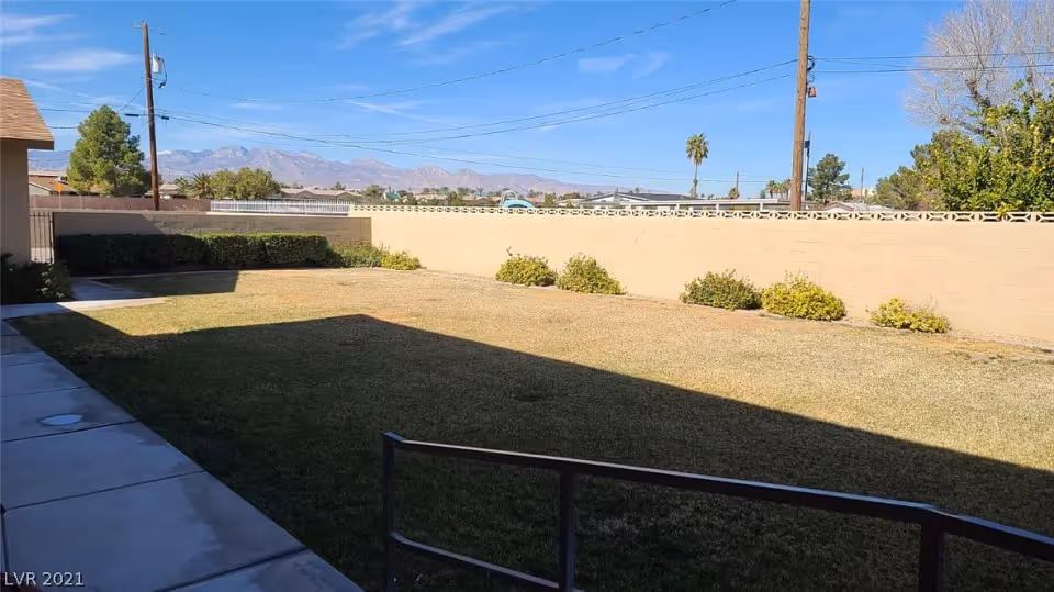 A fenced backyard area with a lawn, some bushes along the perimeter, a concrete walkway on the left side, and a clear blue sky with mountains in the background.