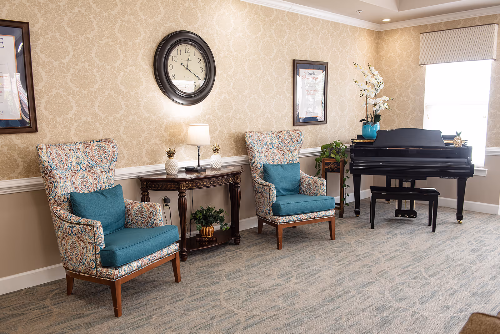 A cozy sitting area in a senior living facility featuring two patterned armchairs with teal cushions, a dark wooden console table with a lamp and decorative pineapples, a large wall clock, framed artwork, a black grand piano with a matching bench, and a window with a patterned valance. The walls have beige damask wallpaper and the floor is carpeted with a subtle pattern.