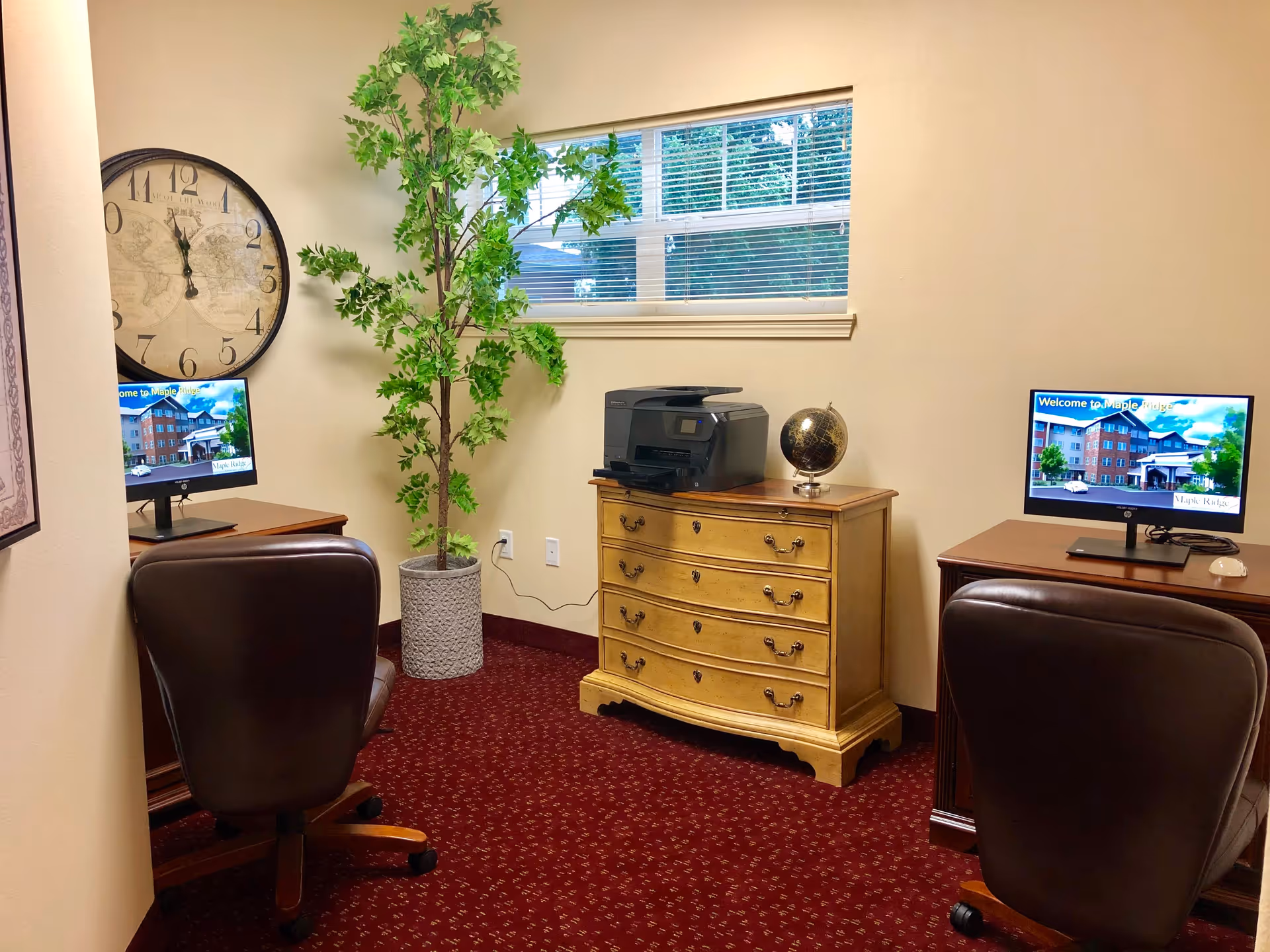 A small office area with two desks each having a computer monitor displaying a welcome message for Maple Ridge. There are two brown leather office chairs in front of the desks. A large wall clock and a tall potted plant are on the left side. A wooden chest of drawers with a printer and a globe on top is positioned against the wall beneath a window with blinds. The room has beige walls and red patterned carpet.