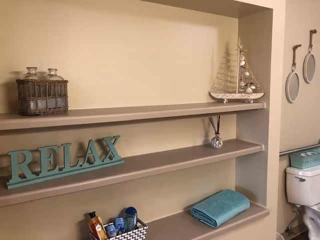 A bathroom shelf with decorative items including a wooden 'RELAX' sign, a small model sailboat, two round hanging mirrors on the wall, a basket with toiletries, and a folded turquoise towel. A toilet is partially visible on the right side of the image.