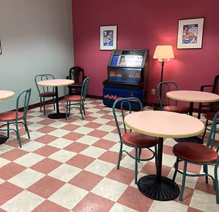 A small dining area with round tables and chairs on a red and white checkered floor. The walls are painted pink and gray, with framed artwork hanging on the pink wall. A floor lamp and a jukebox are also visible against the pink wall.