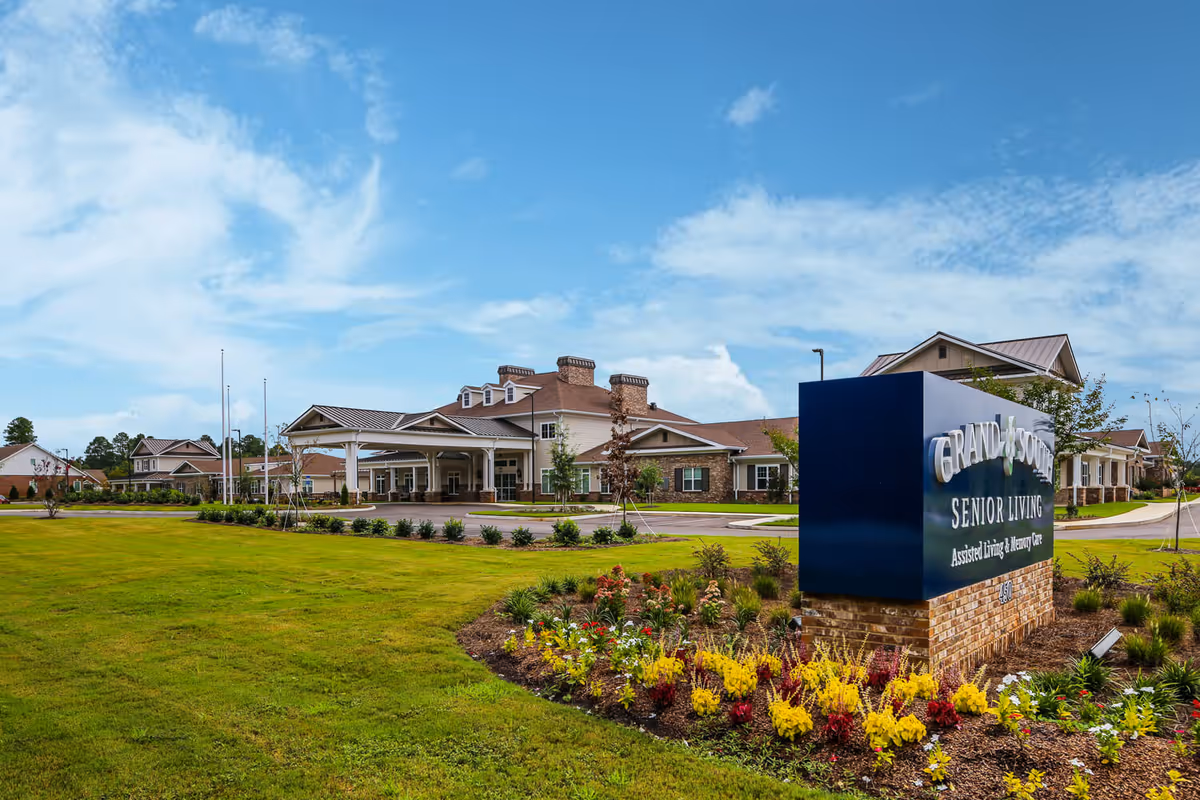 Exterior view of Grand South Senior Living facility with a large blue sign in the foreground surrounded by colorful flowers and a well-maintained lawn. The building has a covered entrance and multiple sections with brick and siding under a partly cloudy sky.
