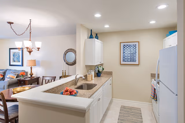 A bright and clean kitchen area with white cabinets, a granite countertop, a sink, and a refrigerator. The kitchen opens to a living room with a gray sofa, a wooden coffee table, a side table with a lamp, and a chandelier overhead. Decorative items include framed artwork on the walls, a bowl of apples on the counter, and vases on top of the cabinets.
