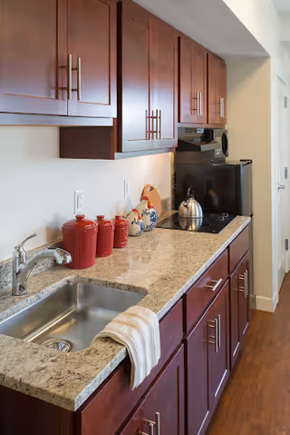 Small kitchenette featuring a granite countertop with a stainless sink, dark wood cabinets, a stovetop with a kettle, microwave, and red canisters.