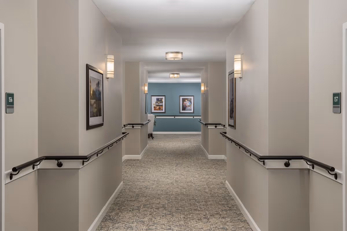 A well-lit hallway in a senior living facility with beige walls, handrails on both sides, framed artwork on the walls, and room number signs 156 and 157 visible. The hallway has a patterned carpet and ceiling lights, leading to a blue accent wall with additional framed pictures.
