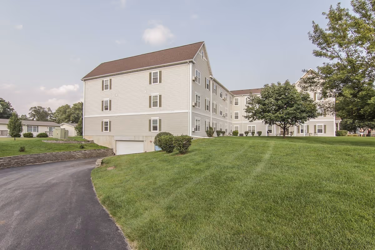 Three-story beige apartment building with a driveway, green lawn, and trees under a partly cloudy sky.