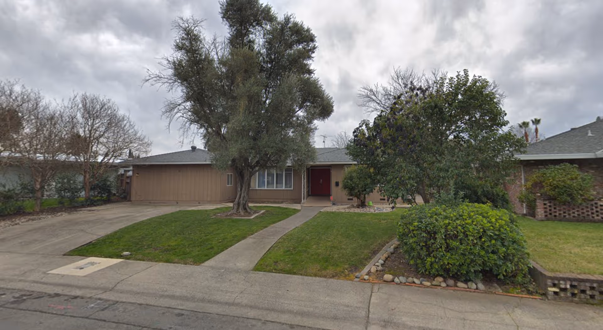 Front exterior view of a single-story residential building with a driveway, a walkway leading to a red front door, and a lawn with trees and bushes under a cloudy sky.