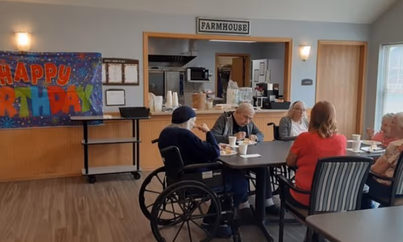 A group of elderly people sitting around a table in a dining area, eating and drinking. One person is in a wheelchair. The room has a 'Happy Birthday' banner on the wall and a sign above a kitchen pass-through window that reads 'FARMHOUSE'.