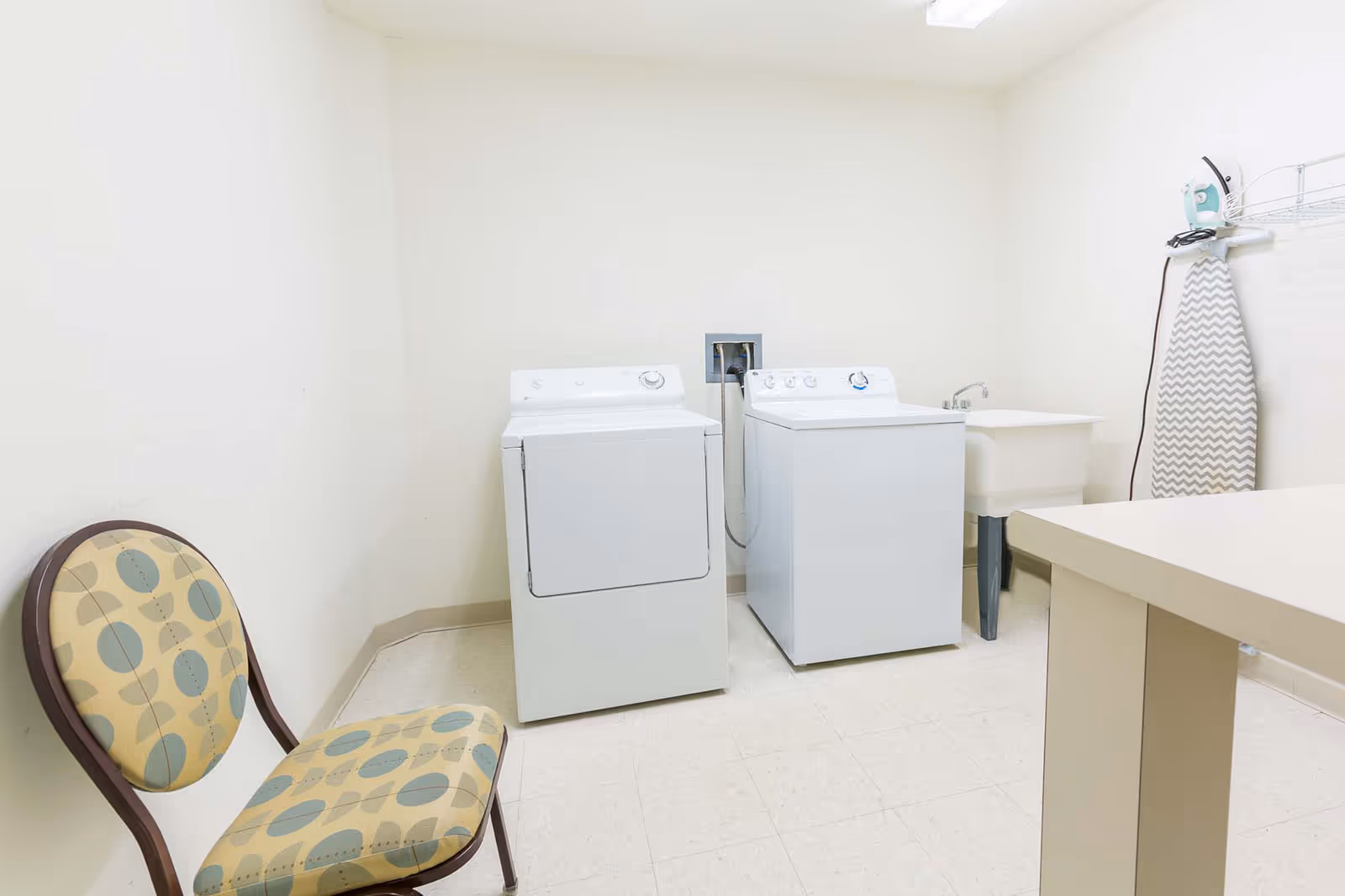 Small laundry room with a washer and dryer, utility sink, ironing board, folding table, and a patterned chair.