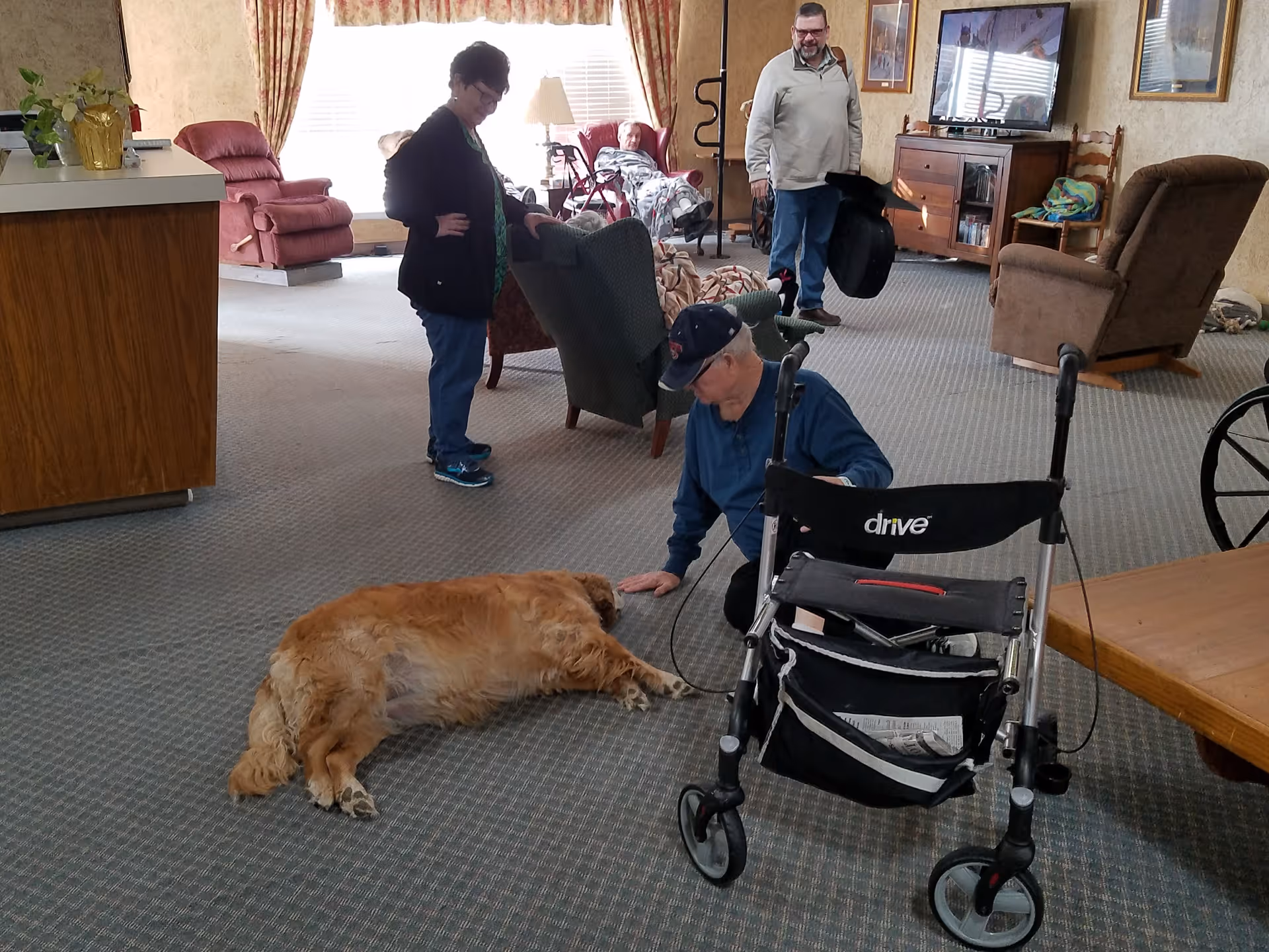 People gathered in a carpeted assisted living common room, with a man kneeling to pet a dog near a walker and several chairs and recliners.