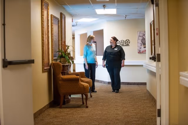 Two women walking and talking in a well-lit hallway of a senior living facility. The hallway has beige walls, carpeted floor, framed artwork, and a cushioned armchair with a plant on a small table beside it.
