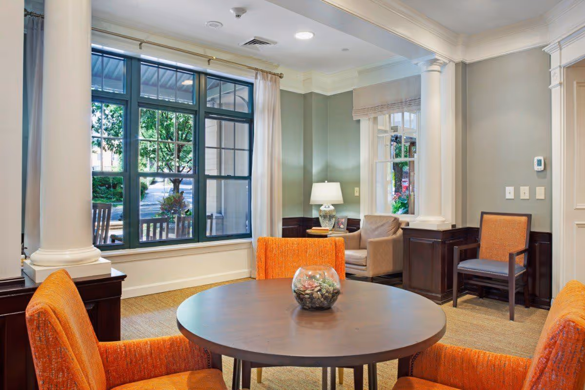 A cozy sitting area in a senior living facility with a round wooden table surrounded by four orange upholstered chairs. There is a glass bowl with decorative stones and plants on the table. The room has large windows with white curtains, allowing natural light to brighten the space. In the background, there is a beige armchair next to a side table with a lamp and framed photo. The walls are painted light gray with white trim and decorative columns.