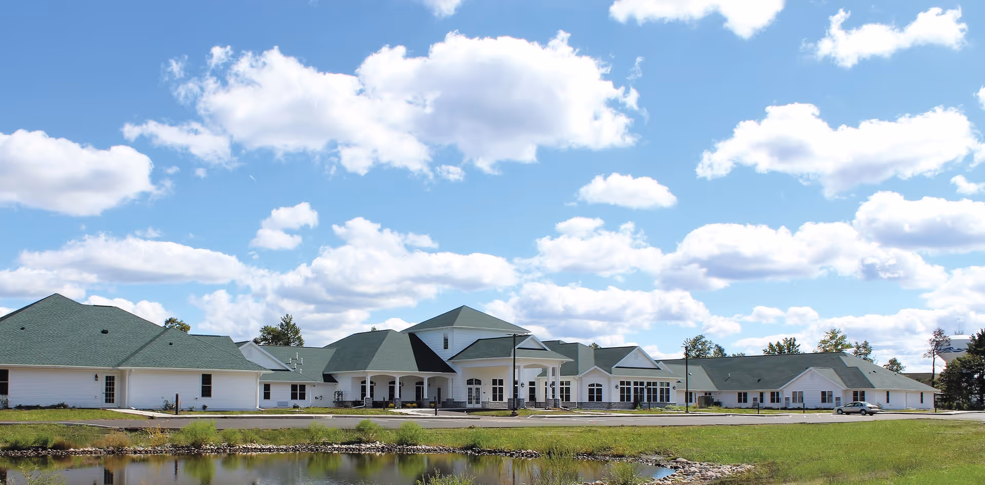 Wide exterior view of a single-story senior living facility with green roofs and white walls, situated near a small pond under a partly cloudy blue sky.