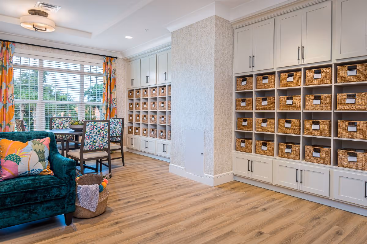 A bright and cozy common area with large windows covered by colorful curtains, a round table with four patterned chairs, a teal velvet armchair with a decorative pillow, and built-in shelves filled with woven baskets for storage. The room has light wood flooring and soft patterned wallpaper.