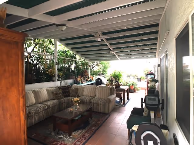 Covered patio with a striped sectional sofa, coffee table, potted plants and exercise equipment under a green-and-white awning.
