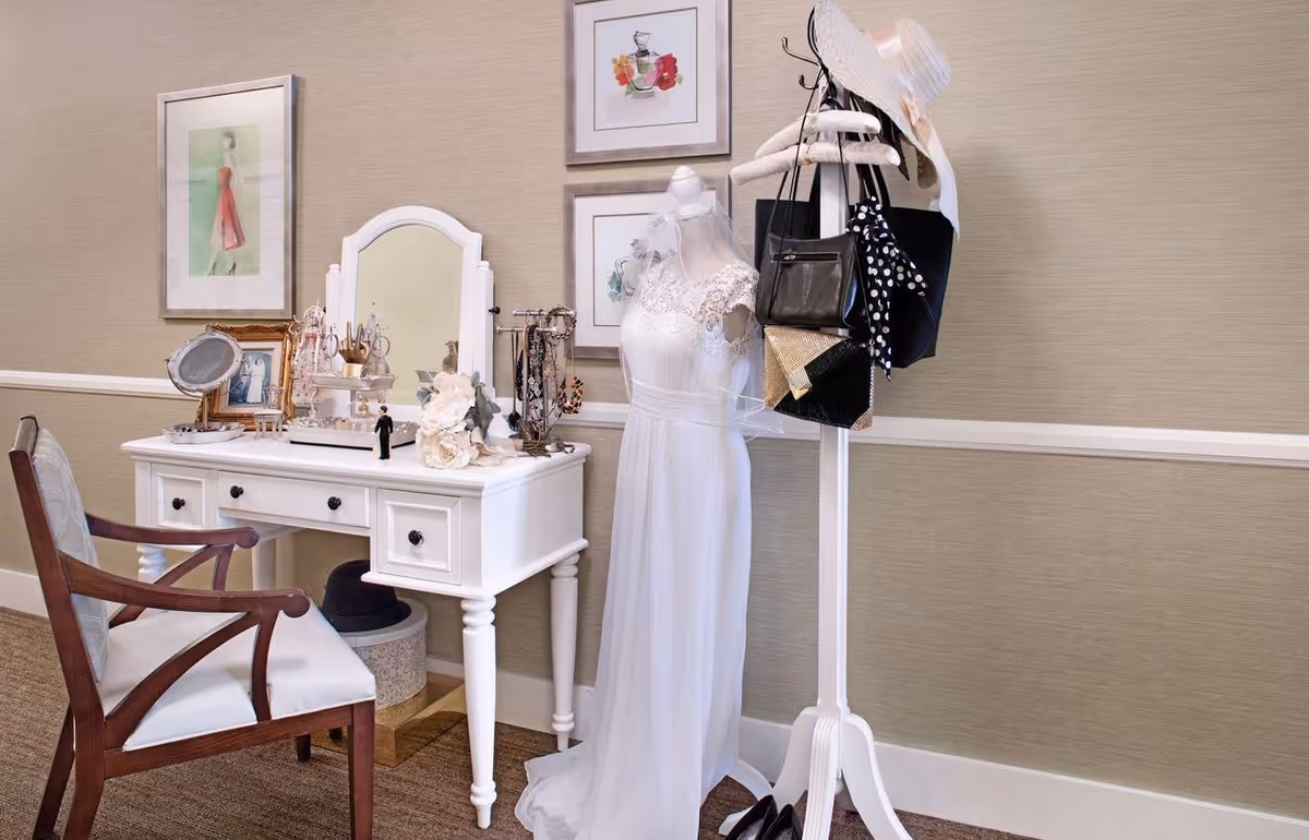 A white vanity table with a mirror, jewelry, and decorative items on top, accompanied by a wooden chair with a white cushion. Next to the vanity is a white mannequin dressed in a white lace wedding gown and veil. A white coat rack holds several handbags, a polka dot scarf, and a sun hat. The background features beige walls with framed artwork.