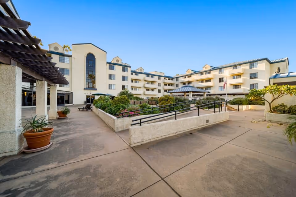 Courtyard and entrance of a multi-story senior living building with landscaped planters, walkways, and balconies.