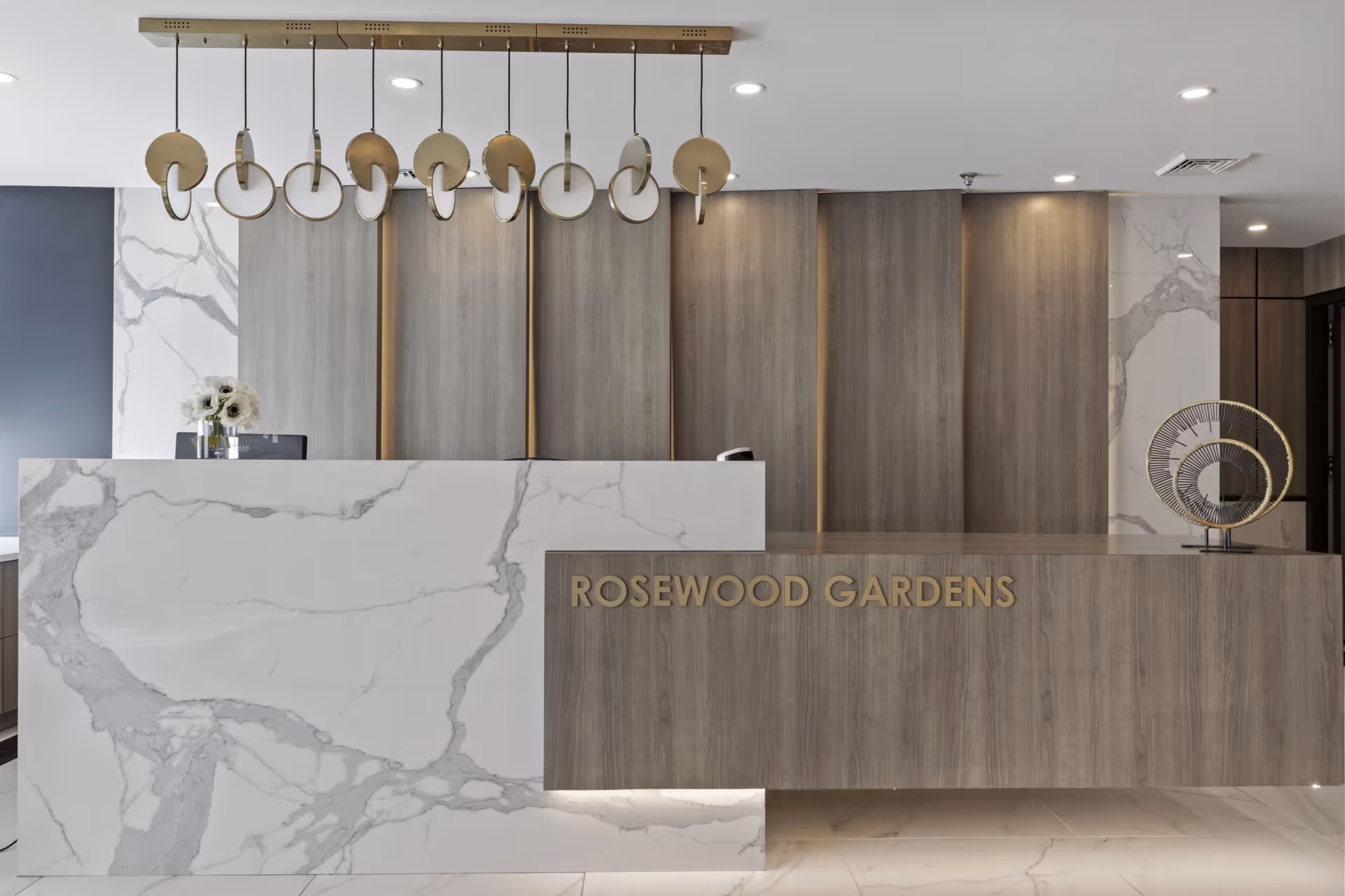 Modern reception desk area with white marble and wood paneling, featuring a decorative hanging light fixture and a sculpture on the counter. The words 'ROSEWOOD GARDENS' are displayed on the wood panel of the desk.