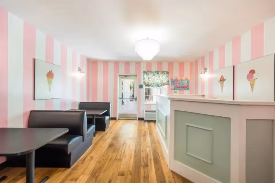 Interior of a small dining area with pink and white striped walls, wooden floor, black booth seating, and tables. There are three framed pictures of ice cream cones on the walls, a chandelier hanging from the ceiling, and a counter with a sign that reads 'Sweet Shoppe'.