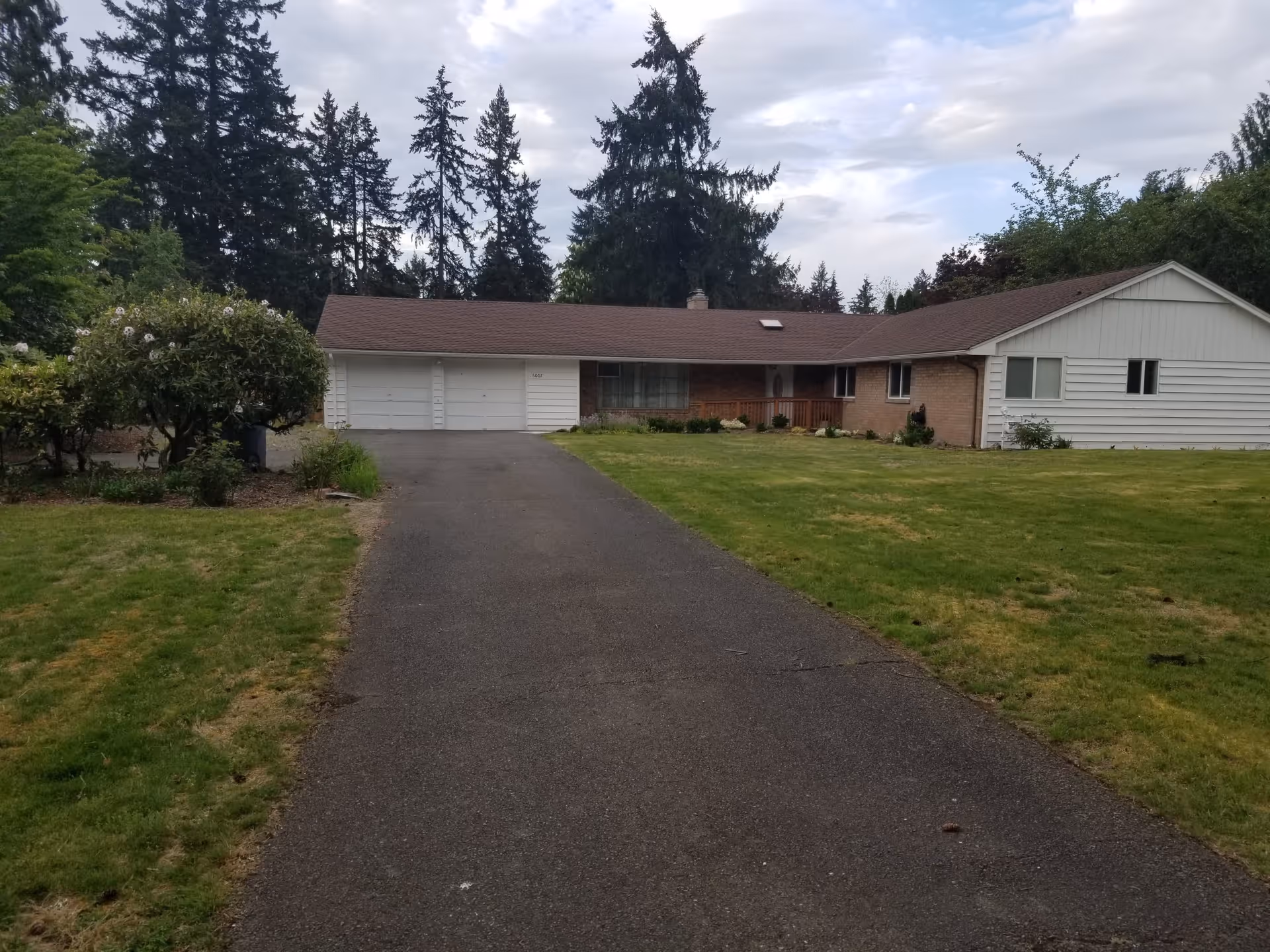 A single-story residential building with a brown roof and white siding, featuring a two-car garage. The house is surrounded by a well-maintained lawn and some bushes, with tall evergreen trees in the background under a partly cloudy sky.
