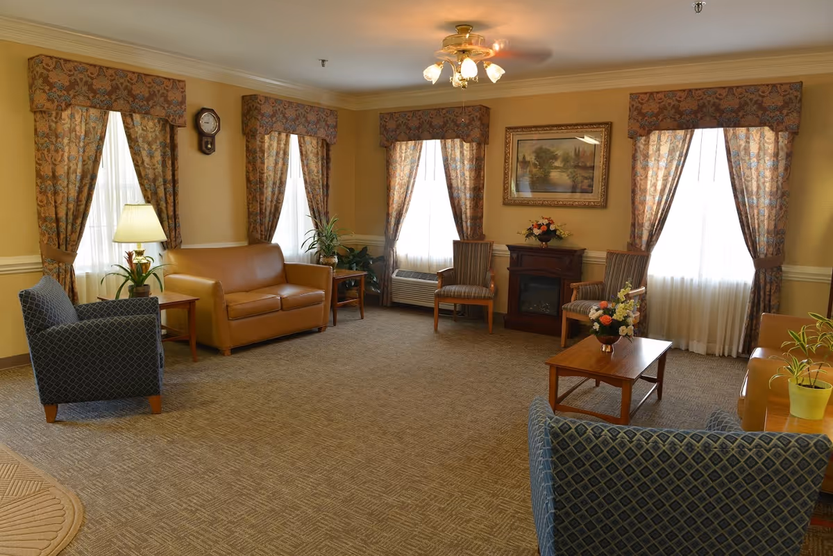 A cozy living room with patterned curtains on four windows, a ceiling fan with lights, a brown leather loveseat, two striped armchairs, two blue patterned armchairs, a wooden coffee table with a flower arrangement, a side table with a lamp and plant, a small electric fireplace with a flower vase on top, and a framed landscape painting on the wall.