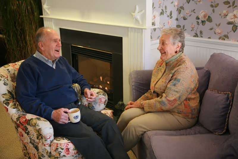 An elderly man and woman sitting in a cozy living room area, engaged in conversation. The man is seated in a floral armchair holding a coffee mug, while the woman sits on a purple couch. Behind them is a fireplace with a small fire burning and floral wallpaper on the wall.