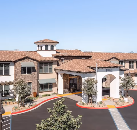 Exterior view of a senior living facility building with a tiled roof, stone and stucco walls, and a covered entrance with an archway. There are small trees and landscaping around the entrance and a clear blue sky above.