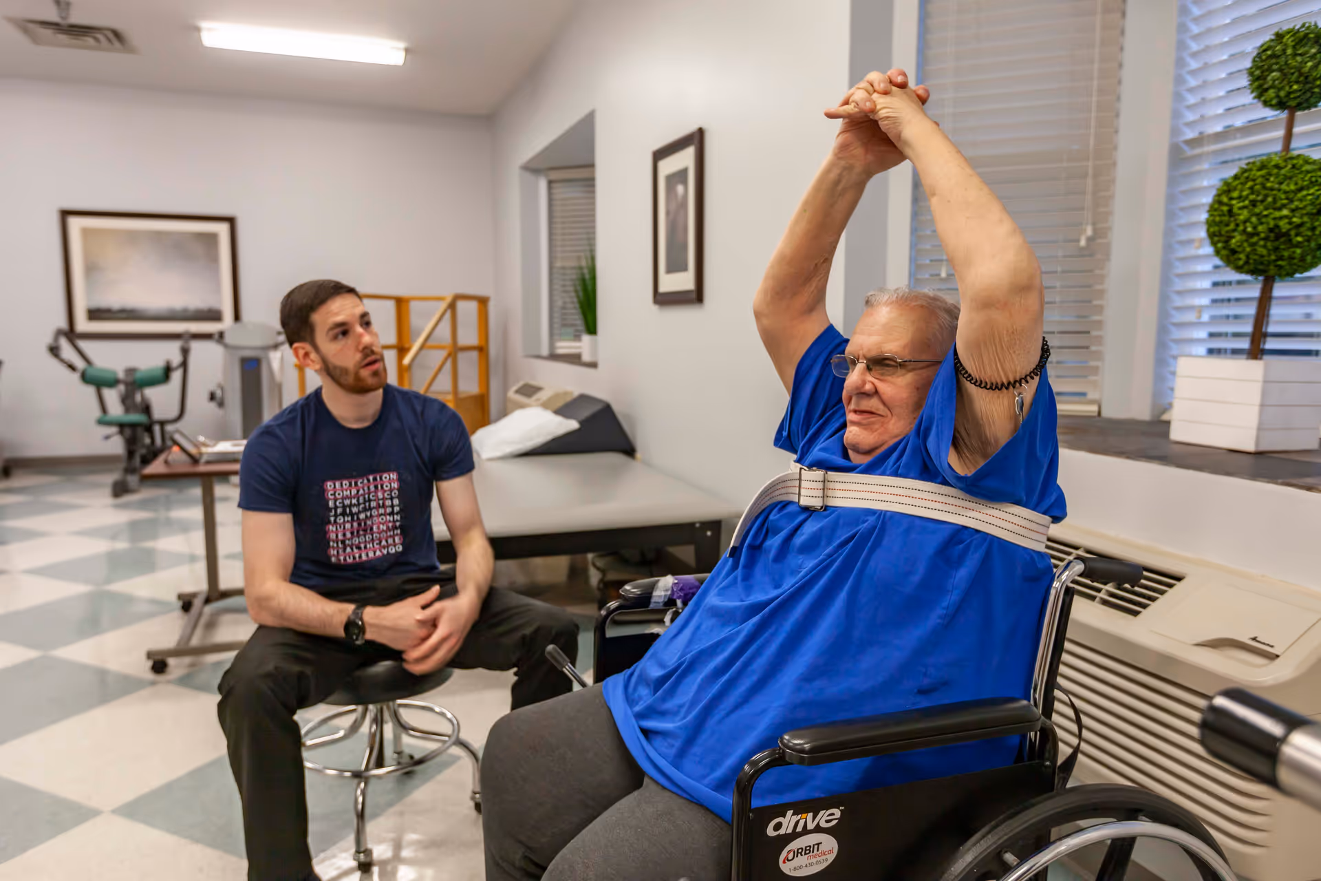 An elderly man in a wheelchair wearing a blue shirt is stretching his arms above his head while a younger man in a dark t-shirt sits nearby observing and supporting him in a rehabilitation room with exercise equipment and framed pictures on the walls.