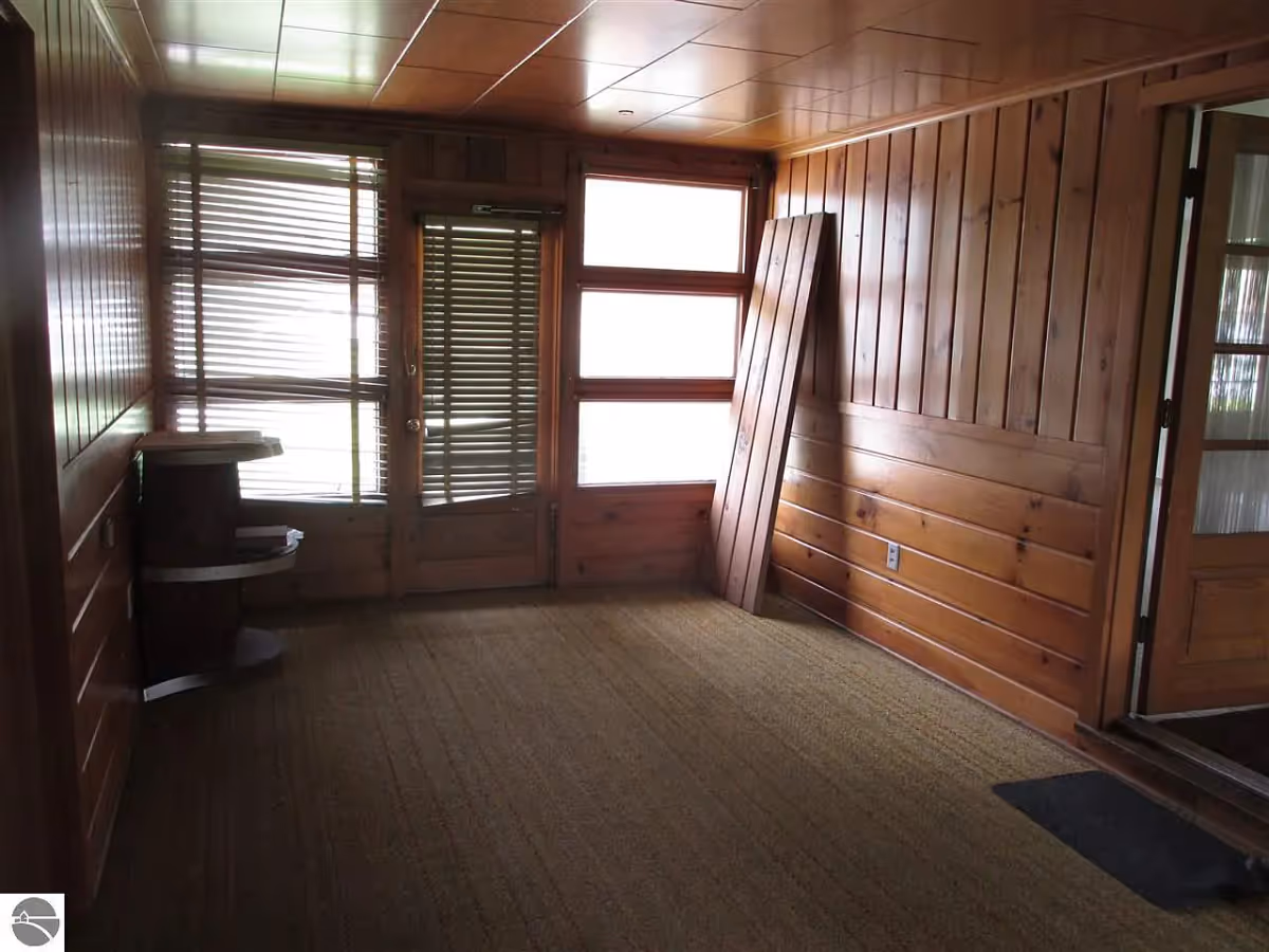 Empty wood-paneled room with carpeted floor, windows with blinds, a door, a small corner shelf and a leaning wooden plank.