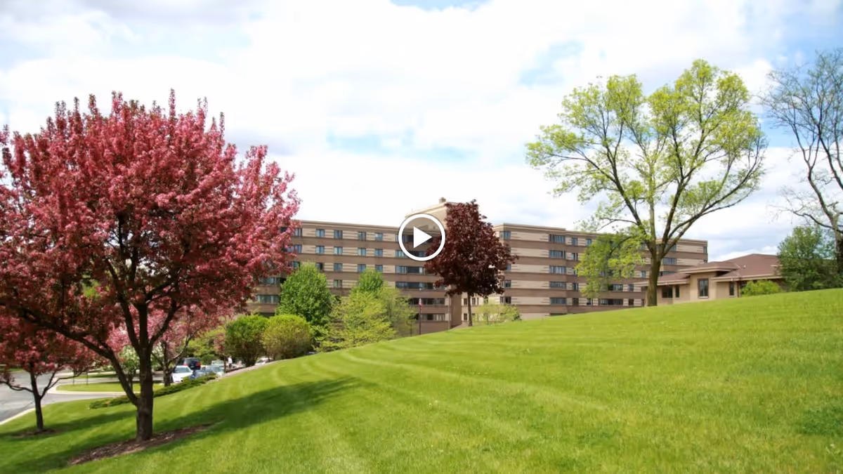 A large senior living facility building in the background with multiple floors and windows, surrounded by a green grassy hill and various trees including a blooming pink tree and green leafy trees under a partly cloudy sky.