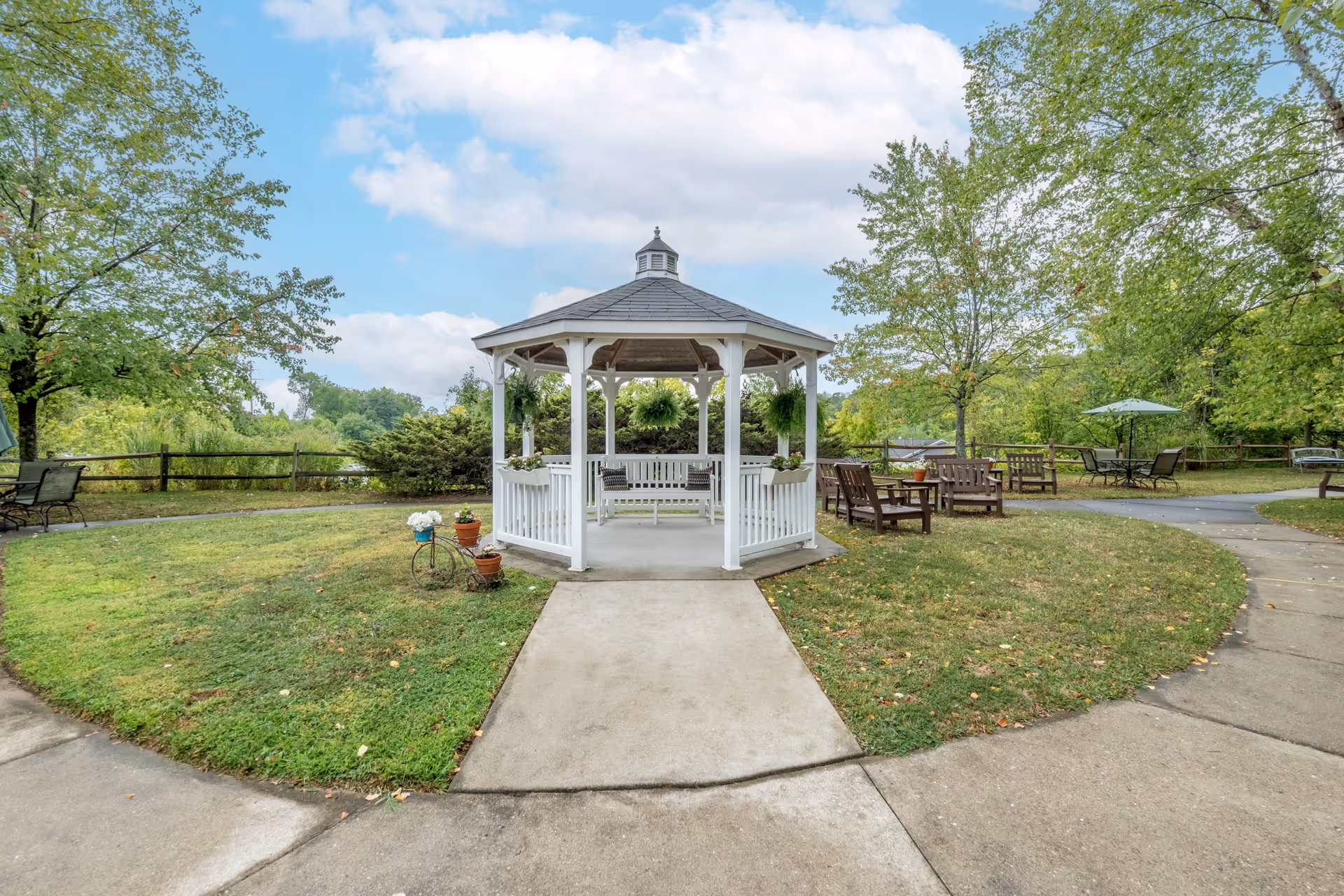 A white gazebo with a bench inside is situated in a grassy outdoor area with paved walkways leading to it. Surrounding the gazebo are trees, wooden benches, a table with an umbrella, and some potted plants. The sky is partly cloudy.