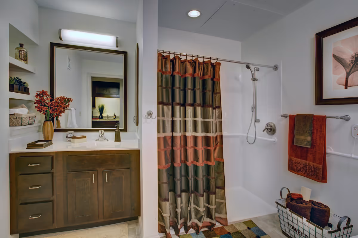 Well-lit bathroom with a wooden vanity and mirror on the left and a shower with a patterned curtain and grab bars on the right.
