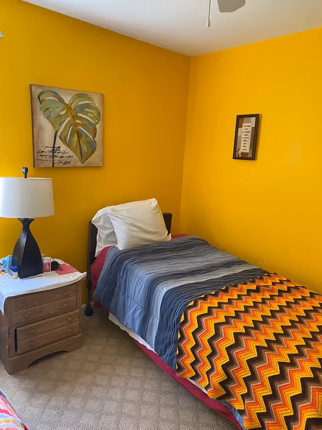 Bright yellow bedroom with a single bed, colorful zigzag blanket, and a wooden nightstand.