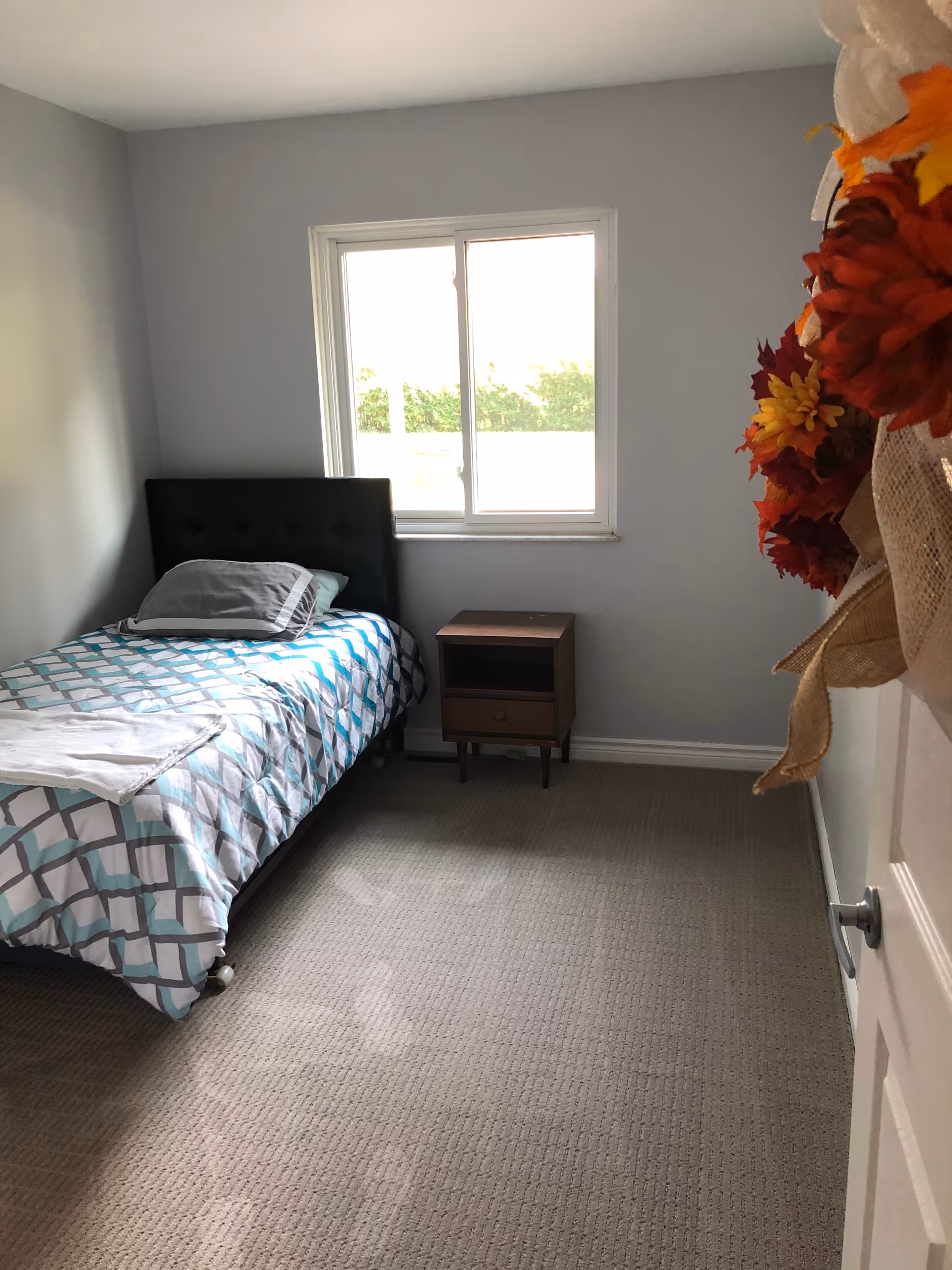A small bedroom with a single bed covered in a geometric patterned bedspread, a pillow, and a folded towel at the foot of the bed. There is a wooden nightstand with a drawer and an open shelf next to the bed. A window with a view of greenery outside is centered on the wall behind the bed. The room has light gray walls and carpeted floor. A door with a fall-themed wreath is partially visible on the right side.