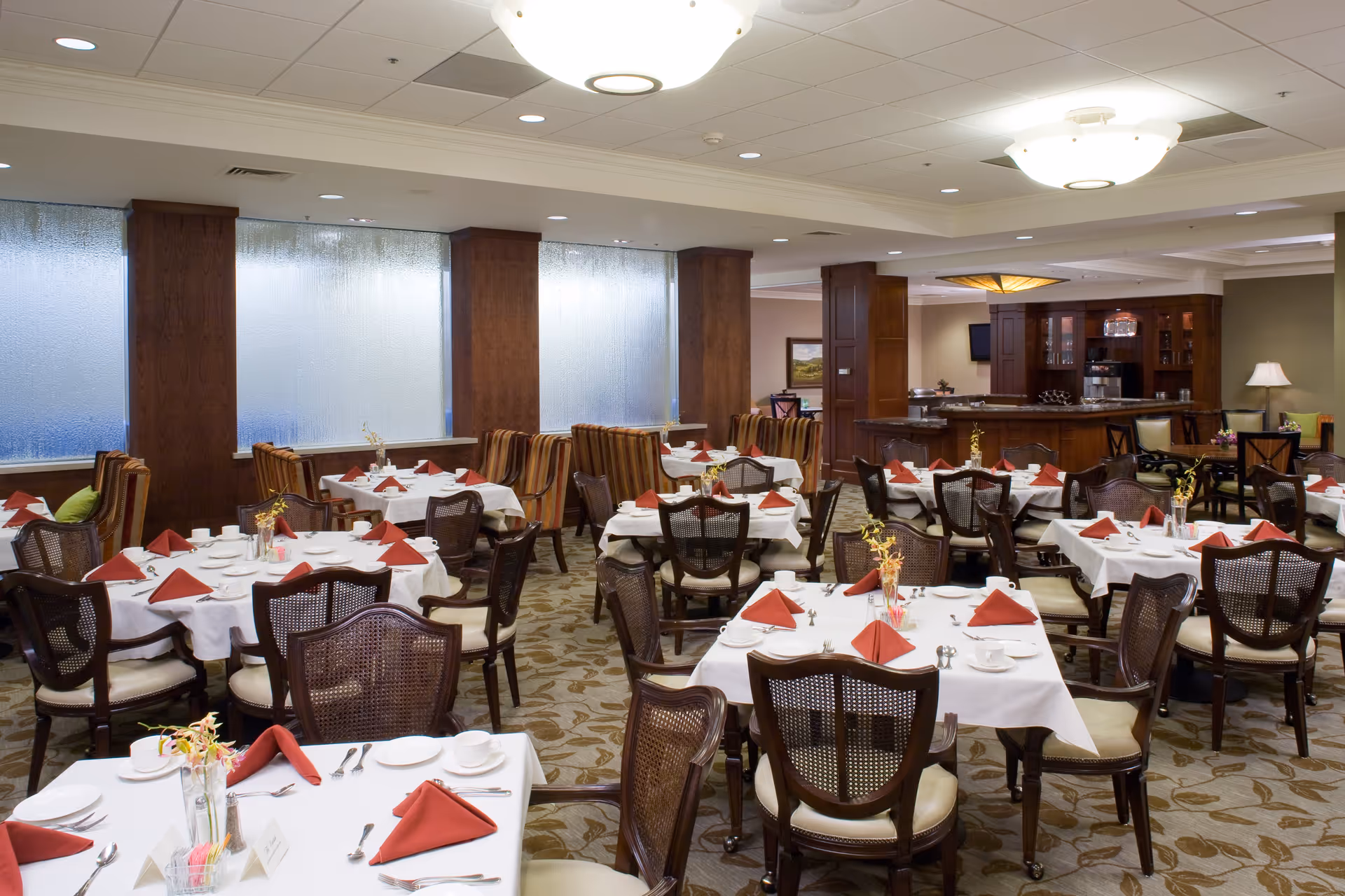 A spacious dining room with multiple tables covered in white tablecloths, each set with red folded napkins, white plates, cups, and silverware. The room features wooden chairs with cushioned seats, patterned carpet, and large frosted windows along one wall. In the background, there is a wooden bar area with cabinets and a coffee machine.