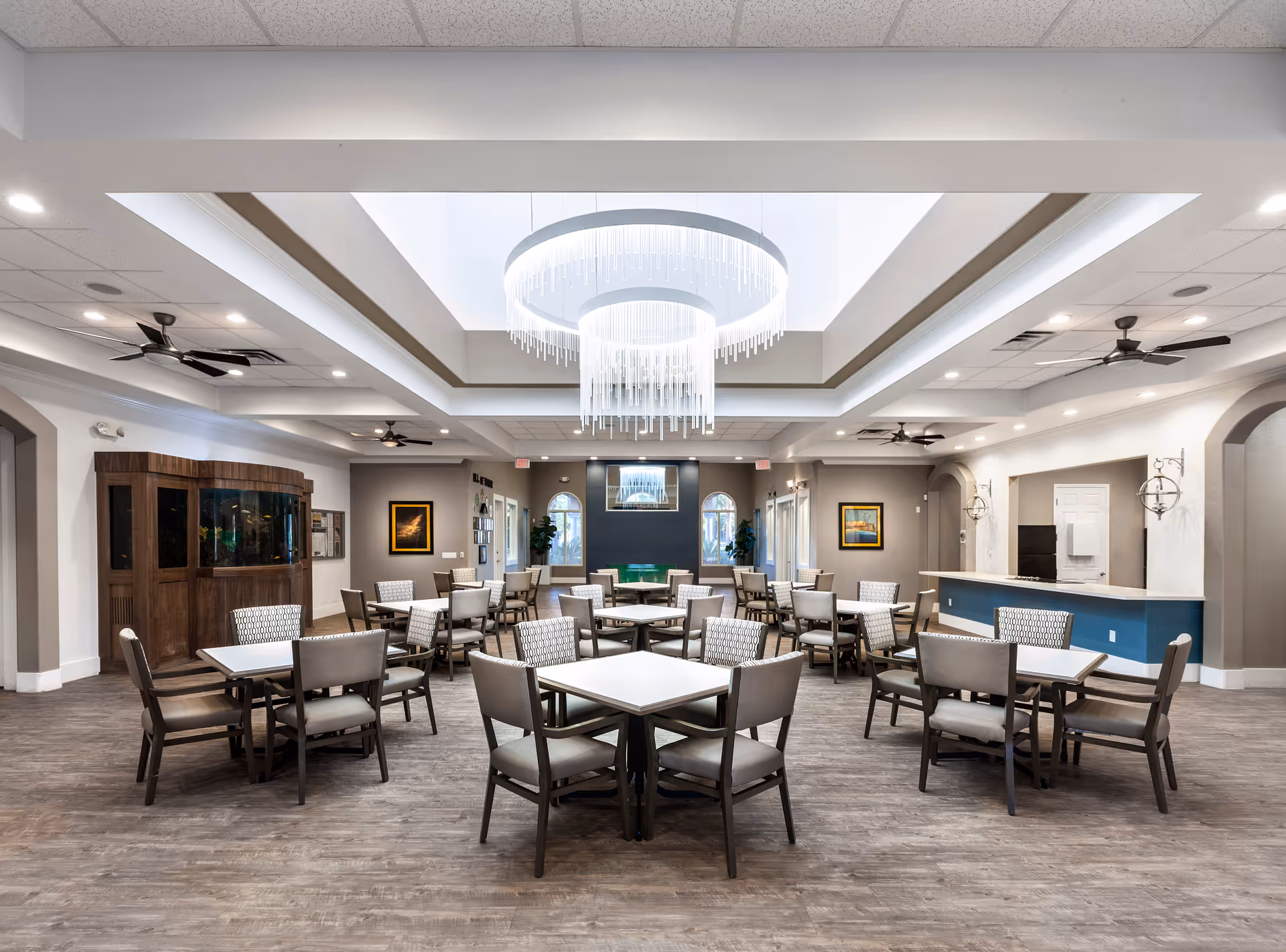 Spacious communal dining room with multiple tables and chairs, a large central chandelier, and a serving counter in a senior living facility.