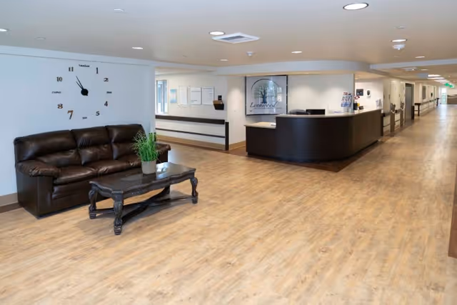 Interior view of a senior living facility hallway with a dark brown leather couch and a wooden coffee table with a potted plant on it. A large wall clock is mounted above the couch. The reception desk is visible in the background with the facility name Lynnwood displayed behind it. The hallway extends to the right with doors along the corridor.