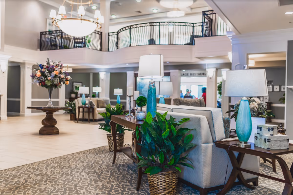 Spacious and well-lit senior living facility lobby with comfortable seating areas, decorative plants, table lamps with blue bases, a large floral arrangement on a round wooden table, and a second-floor balcony overlooking the area.