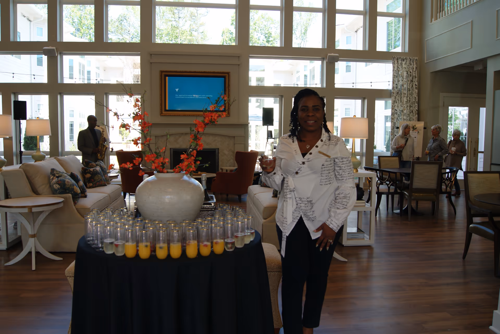 A woman standing in a spacious, well-lit living room holding a glass of beverage. In front of her is a round table covered with a black cloth, holding many glasses filled with orange juice and another clear drink, along with a large vase with orange flowers. In the background, a man is playing a saxophone near a beige sofa, and three elderly women are conversing near a dining table. The room has large windows letting in natural light and modern decor.