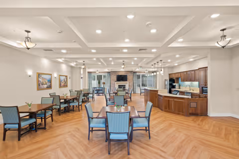Spacious dining room with multiple wooden tables and blue cushioned chairs arranged neatly. The room features a wooden floor with a herringbone pattern, a kitchen area with wooden cabinets and a countertop on the right, and a fireplace with a TV mounted above it at the far end. The walls are decorated with framed paintings, and the ceiling has recessed lighting and hanging light fixtures.