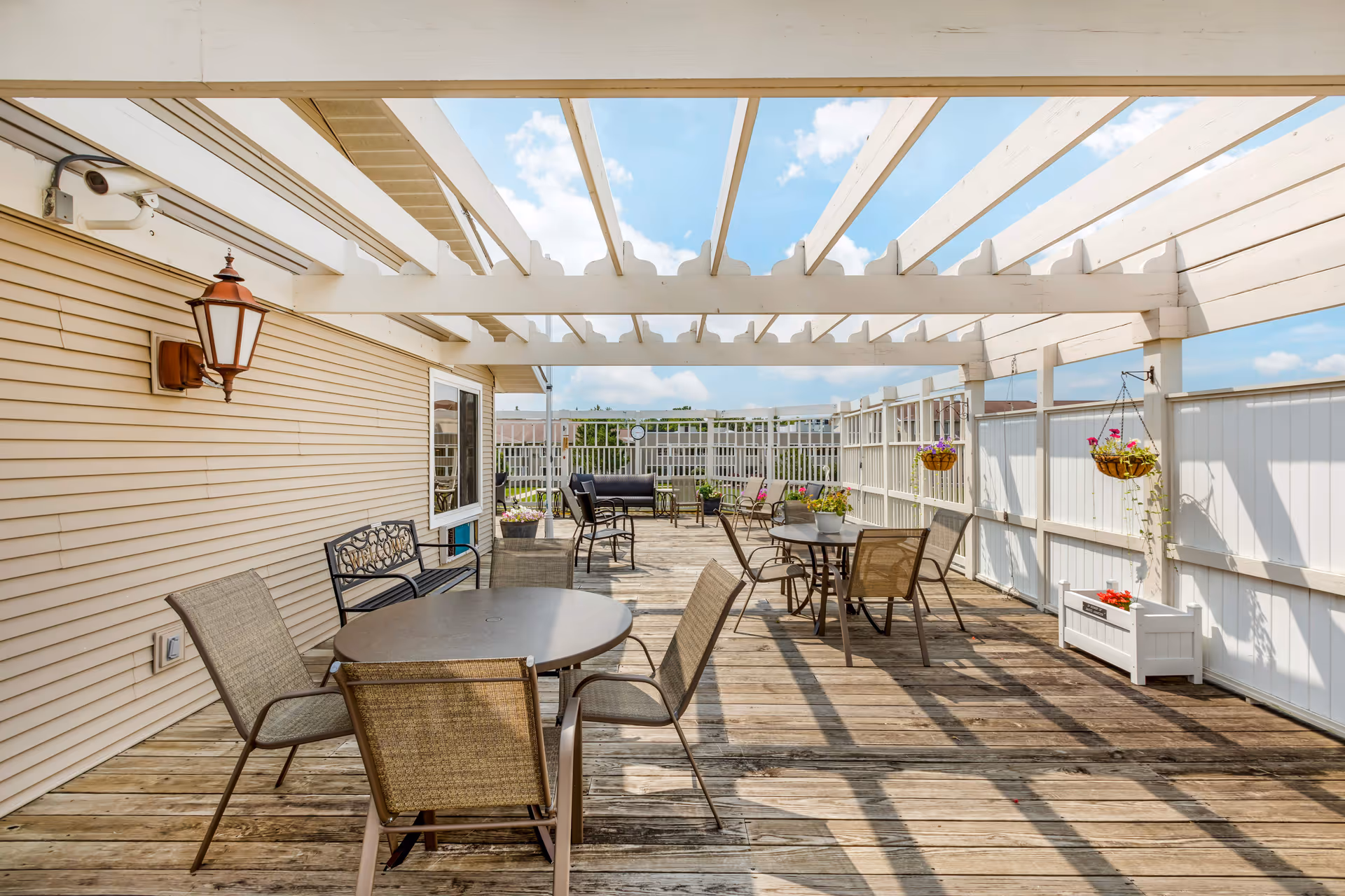 Outdoor patio area with wooden deck flooring and white pergola overhead. Several round tables with chairs are arranged along the patio, with hanging flower baskets and potted plants adding decoration. The patio is enclosed by a white fence and has a beige siding wall with a lantern-style light fixture.