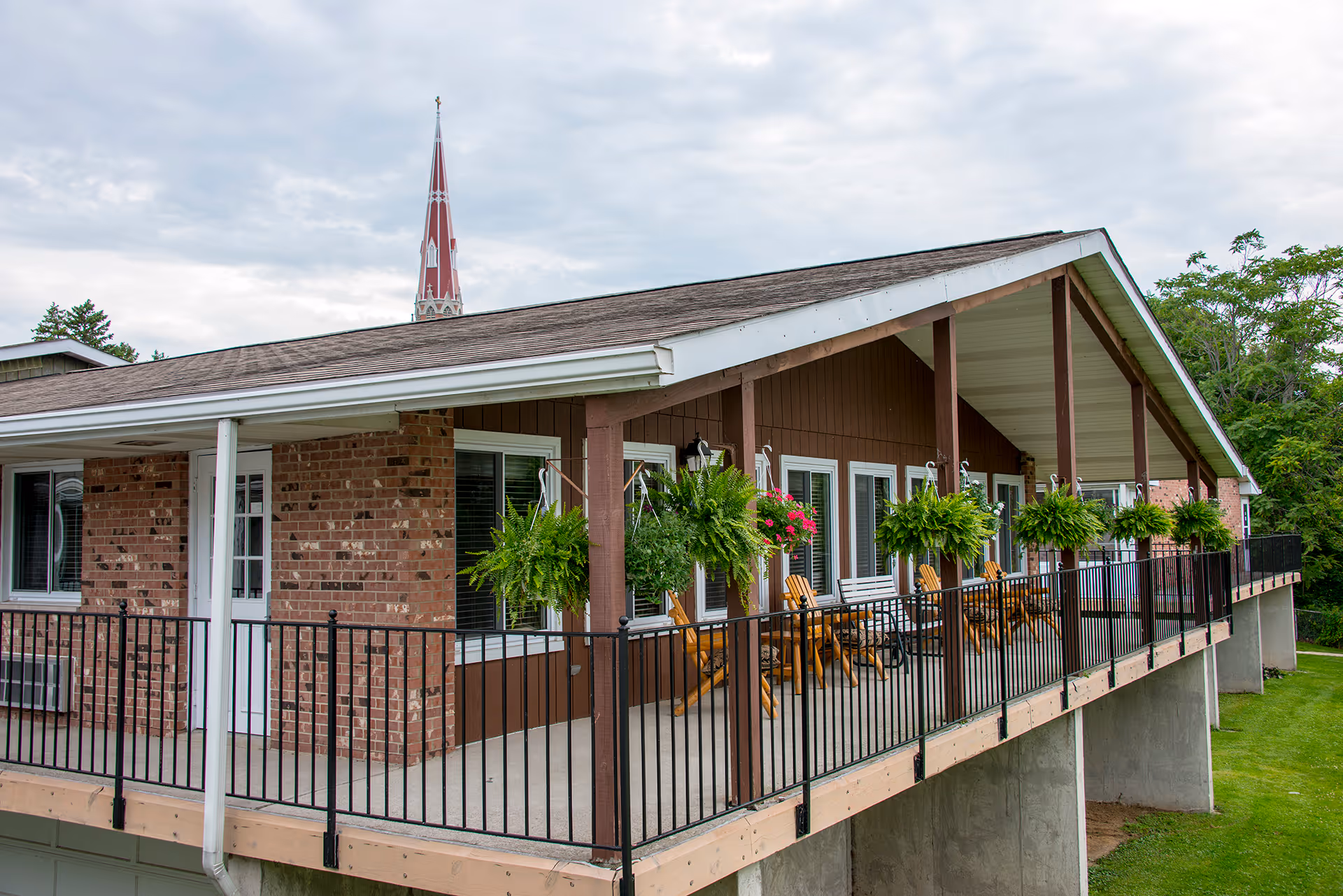 Exterior view of a senior living facility balcony with hanging plants and wooden chairs, featuring a brick and wood facade under a cloudy sky with a church steeple visible in the background.
