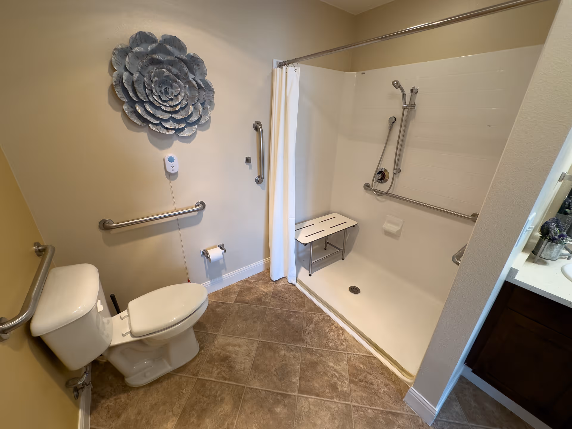 Accessible bathroom with a toilet, multiple grab bars, a walk-in shower with a bench and a large metal flower wall decoration.