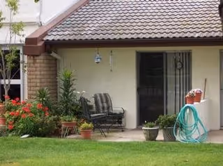 A covered patio area outside a residential building with two cushioned chairs, several potted plants, a garden hose coiled on a holder, and a well-maintained lawn in the foreground.