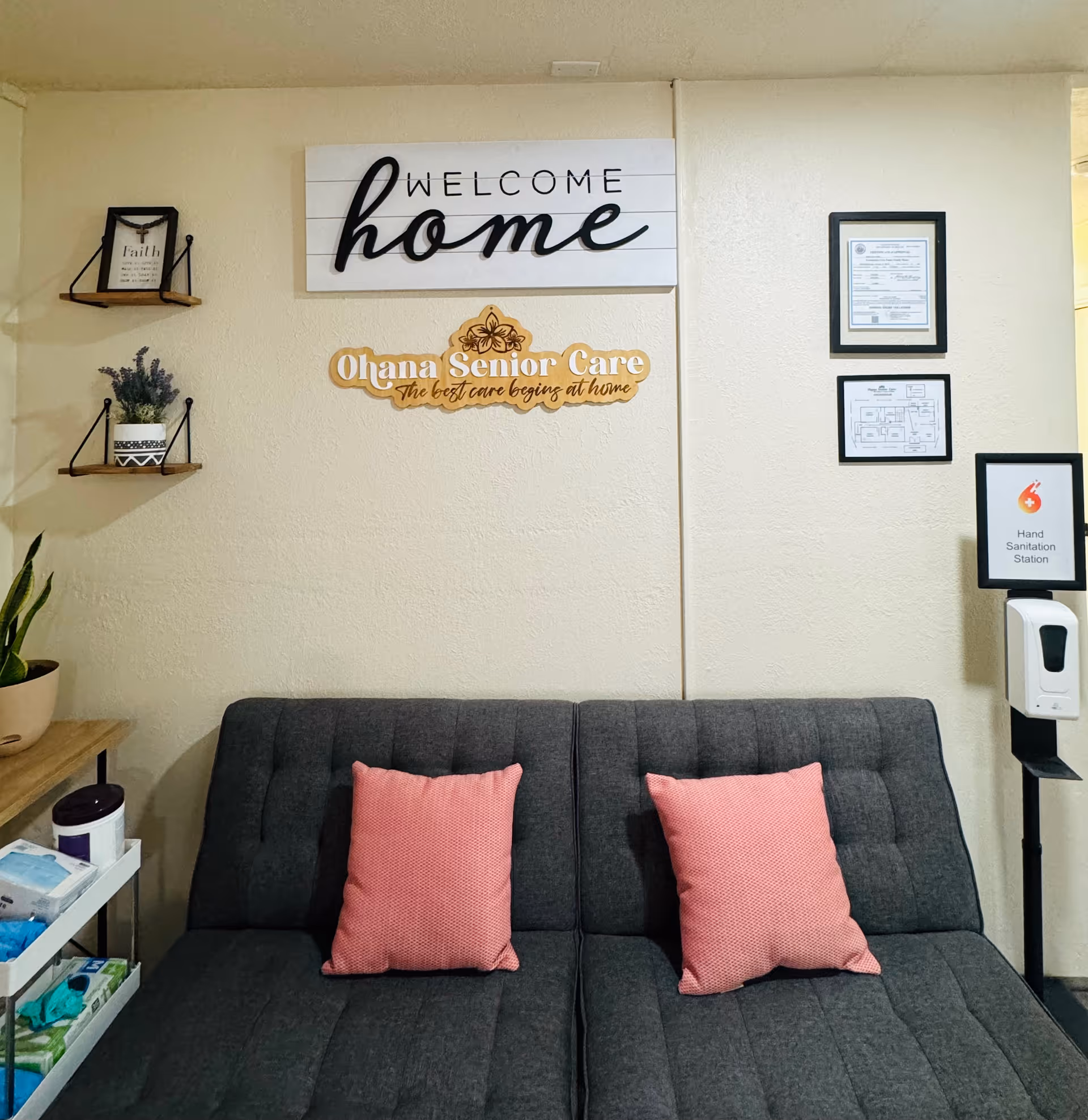 Interior view of a senior care facility waiting or common area with a dark gray cushioned bench featuring two pink pillows. Above the bench, there is a wall decoration with the words 'WELCOME home' and a sign that reads 'Ohana Senior Care The best care begins at home.' On the left side, there are two small wall shelves with a framed quote and a potted plant. On the right side, there are framed certificates and a hand sanitation station with a dispenser.