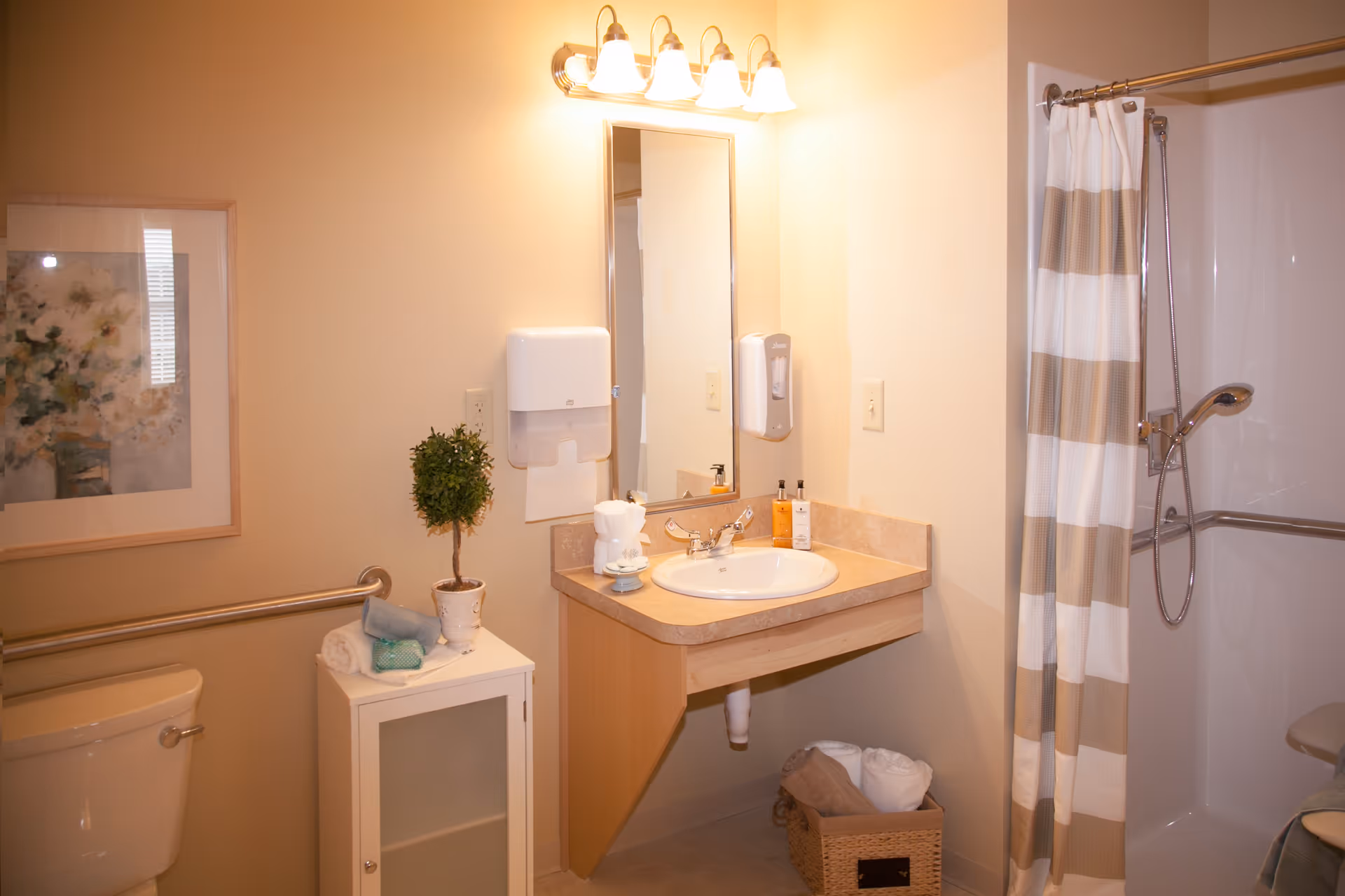 A clean and well-lit bathroom featuring a toilet with a grab bar, a small cabinet with folded towels and a potted plant, a sink with a mirror above it, soap dispensers, and a shower area with a striped shower curtain.